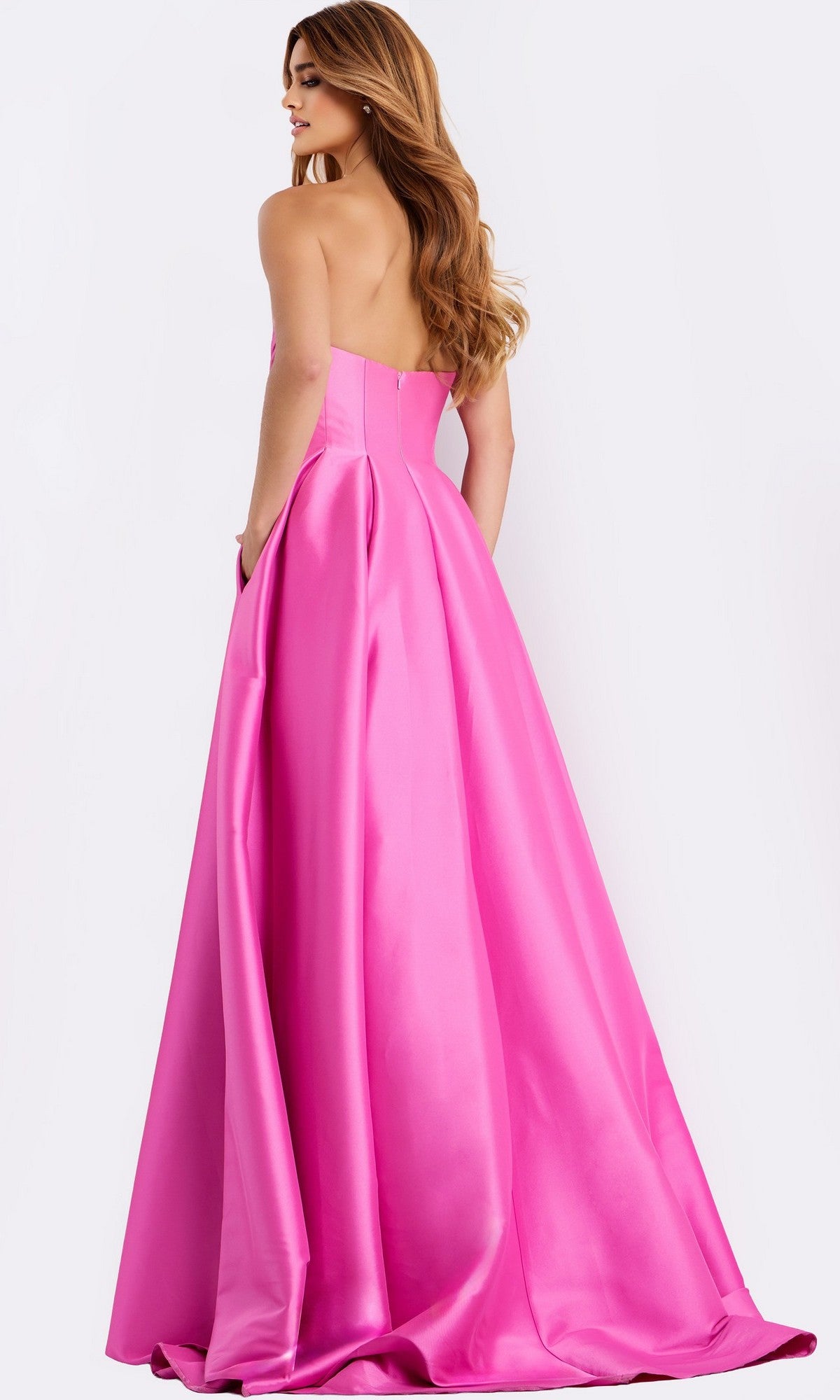 A woman models the Jovani 47570 Strapless Long Pleated Prom Dress, featuring a bright pink a-line silhouette and flowing flared skirt, standing with her back turned as the gown cascades to the floor.